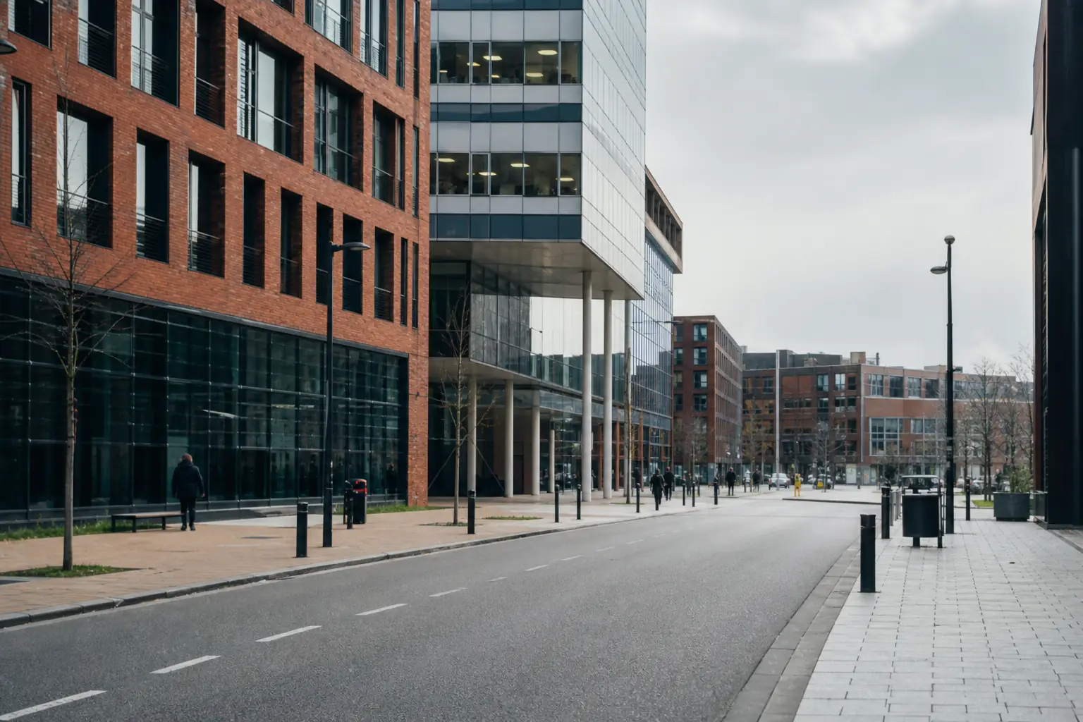 Rue d’un quartier d’affaires du Nord : immeubles de bureaux en verre mêlés à des façades de brique rouge, ciel couvert